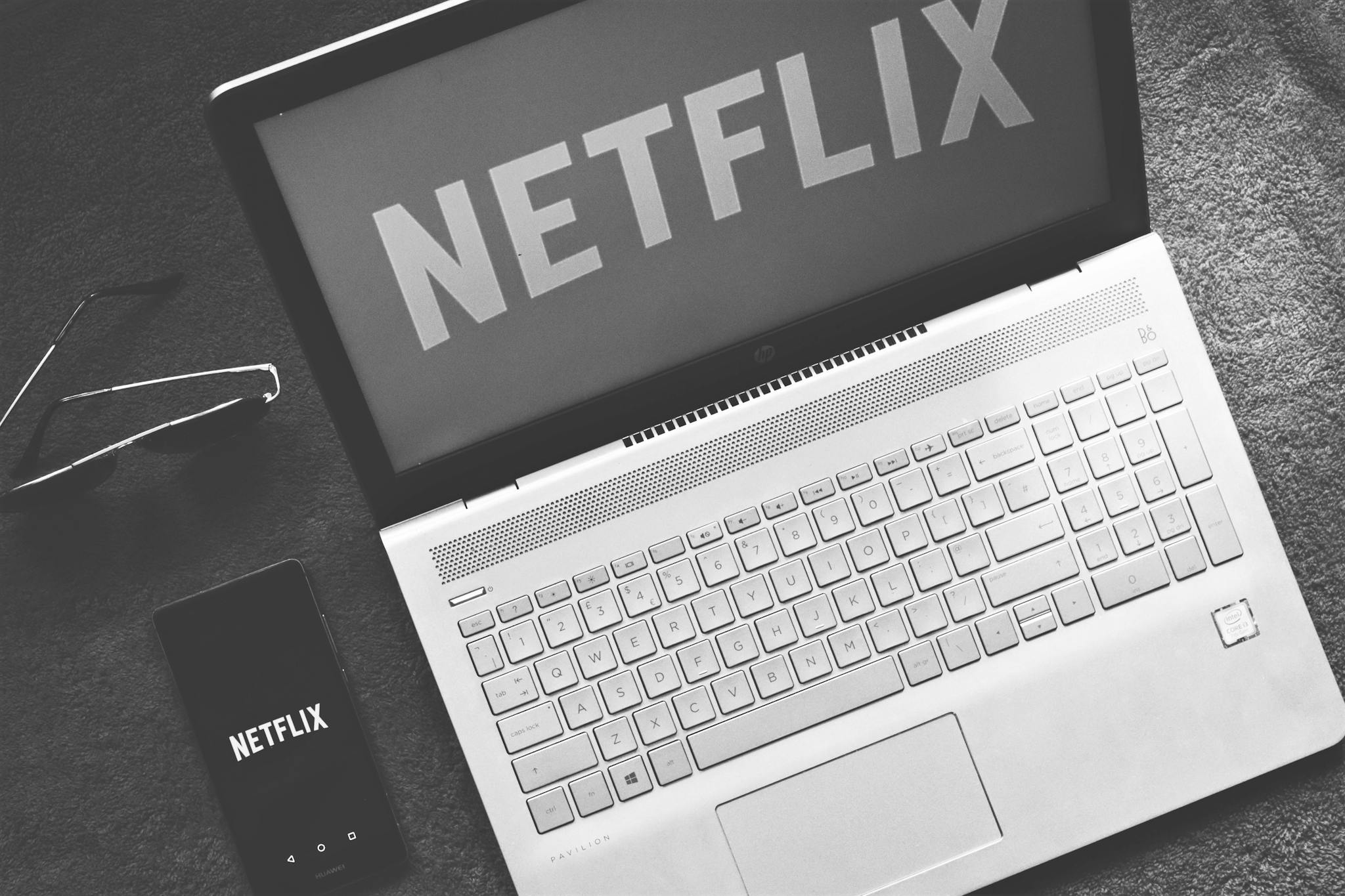 Monochrome image showcasing a laptop with Netflix on screen, smartphone, and sunglasses.